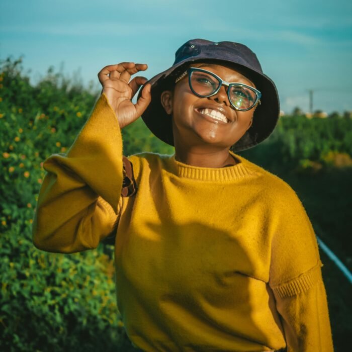 Cheerful woman in glasses and bucket hat smiling in a sunny outdoor setting.